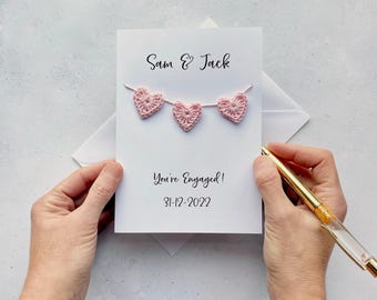 Może przedstawiać: Biała kartka zaręczynowa z imionami "Sam & Jack" i wiadomością "You're Engaged!" wraz z datą 31-12-2022. Karta zawiera sznur trzech różowych serc szydełkowych. Złoty długopis jest trzymany w pobliżu karty.