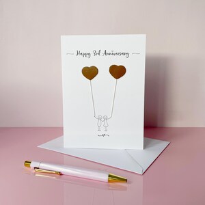 May include: A white anniversary card with the text "Happy 3rd Anniversary" and two gold heart-shaped balloons. The card features a simple illustration of two stick figures holding hands. A white envelope and a pink and gold pen are also in the image.