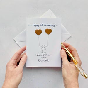 May include: A white anniversary card with the text "Happy 3rd Anniversary." Two brown heart-shaped balloons are held by stick figures. The names "Emma & Robin" and the date "22-08-2020" are also printed on the card.