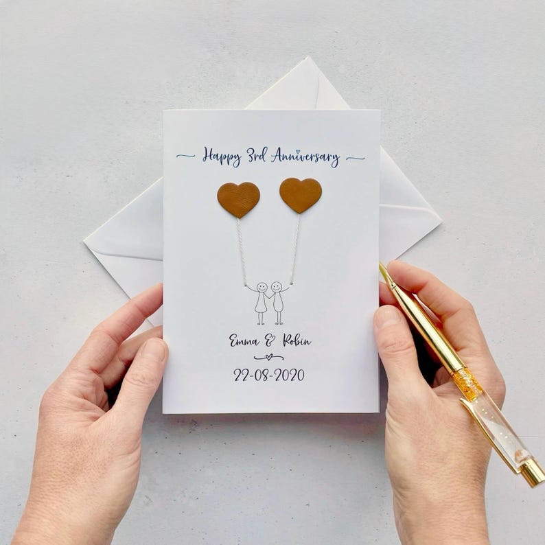 A white card featuring a simple line drawn couple, each are holding a heart shaped balloon made from genuine leather. Happy 3rd anniversary is printed at the top of the card and the couples name and wedding date are printed at the bottom.