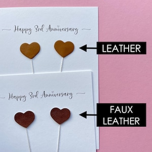 May include: Two white cards with the text "Happy 3rd Anniversary" and heart-shaped decorations. The top card has two tan leather hearts, and the bottom card has two faux leather hearts in a darker brown color.