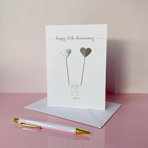 Op de afbeelding: Een witte jubileumkaart met de tekst "Happy 10th Anniversary" en twee hartvormige ballonnen. De ballonnen worden vastgehouden door stokfiguren. Een witte envelop en een witte en gouden pen staan op de voorgrond.