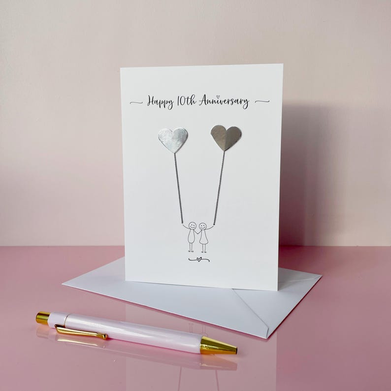 Op de afbeelding: Een witte jubileumkaart met de tekst "Happy 10th Anniversary" en twee hartvormige ballonnen. De ballonnen worden vastgehouden door stokfiguren. Een witte envelop en een witte en gouden pen staan op de voorgrond.