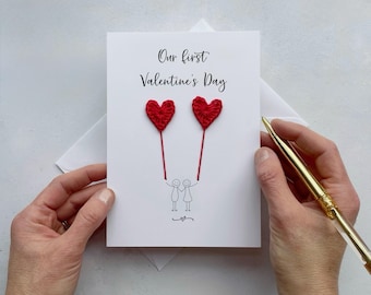 May include: A white greeting card with the text Our first Valentine's Day in black script. Below the text are two red crocheted heart balloons attached to thin red strings held by two simple black line-drawn stick figures holding hands.