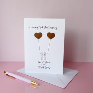 May include: A white anniversary card with the text "Happy 3rd Anniversary" and the names "Jim & Shona" with the date "03-09-2022". The card features two brown heart-shaped balloons held by stick figures. A pink pen with a gold tip rests on the surface.