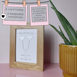 May include: A framed anniversary print with a light wood frame. The print features two heart-shaped balloons connected to stick figures, with the text "Happy 10th Anniversary", names, and a date. The image includes text that reads "A card today... A keepsake tomorrow."