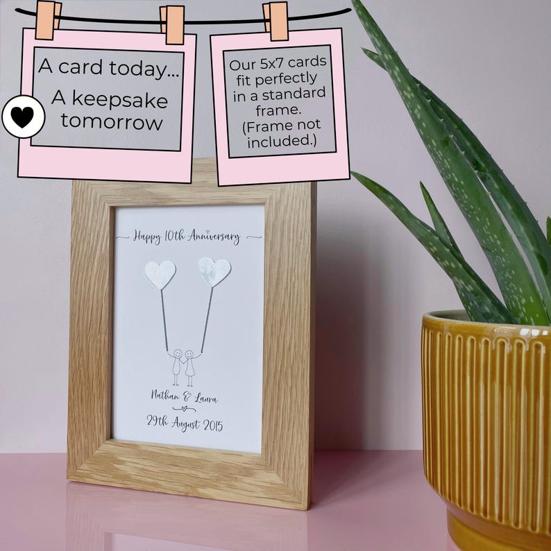 May include: A framed anniversary print with a light wood frame. The print features two heart-shaped balloons connected to stick figures, with the text "Happy 10th Anniversary", names, and a date. The image includes text that reads "A card today... A keepsake tomorrow."