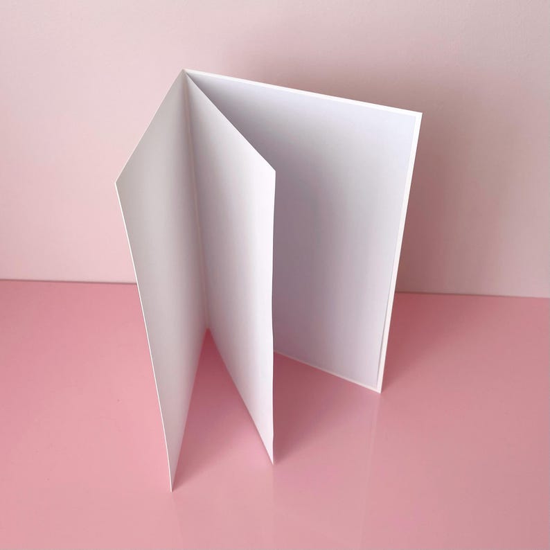 Op de afbeelding: Een witte, gevouwen kaart staat open op een roze oppervlak. De kaart is gemaakt van dik papier en heeft een strak, minimalistisch ontwerp. De achtergrond is zachtroze, wat een eenvoudige en elegante compositie cre&euml;ert.