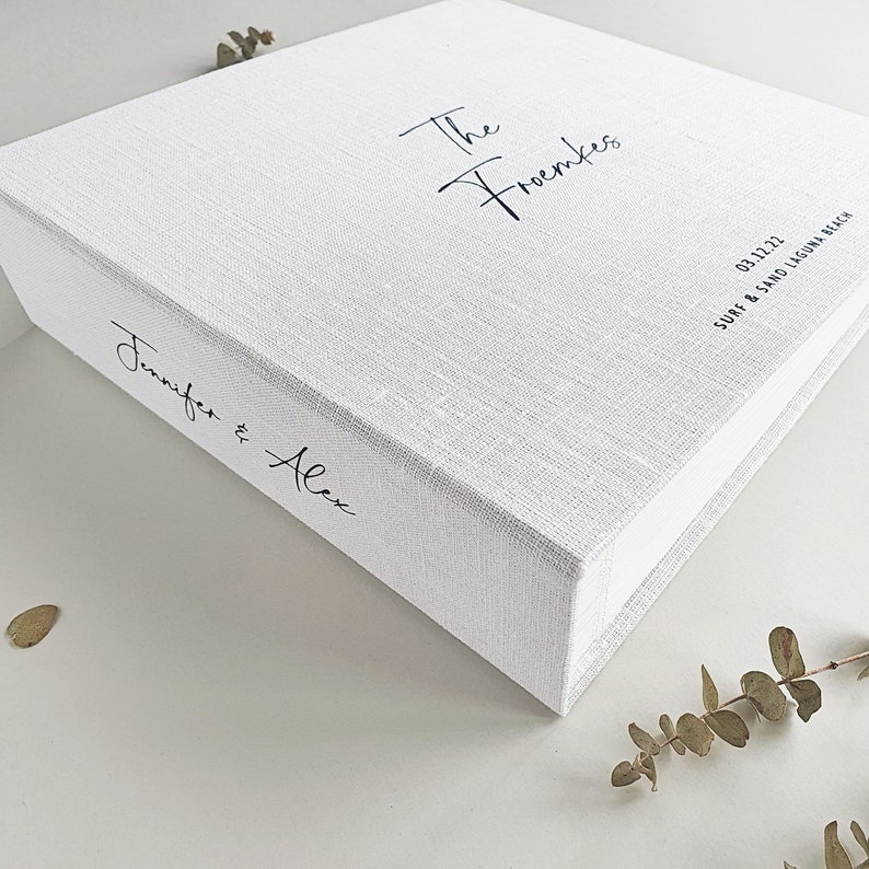 May include: A white linen wedding album with the text 'The Froemkes' and 'Jennifer & Alex' printed in black. The date '03.12.22' and the text 'SURF & SAND LAGUNA BEACH' are also printed in black on the album.