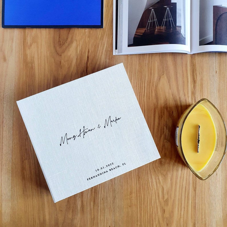 May include: A white linen-covered photo album with the text "Mang & Huan & Marfa" and the date "10.01.2022" and the location "Fernandina Beach, FL" printed on the front.