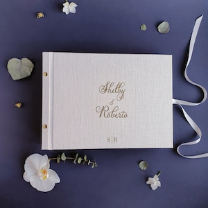 Könnte beinhalten: Ein weißes Leinen-Hochzeitsalbum mit goldenen Lettern, auf dem "Shelby & Roberto" und "S|R" steht. Das Album ist mit einem weißen Band gebunden.