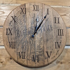 May include: A round wooden clock with Roman numerals. The clock face is made of light brown wood with dark brown markings. The clock hands are black. The clock is mounted on a wooden wall.