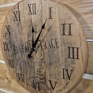 May include: A round wooden clock with Roman numerals and black clock hands. The clock face is made from reclaimed wood and features the text "BUFFALO TRACE". The clock is mounted on a wooden wall.