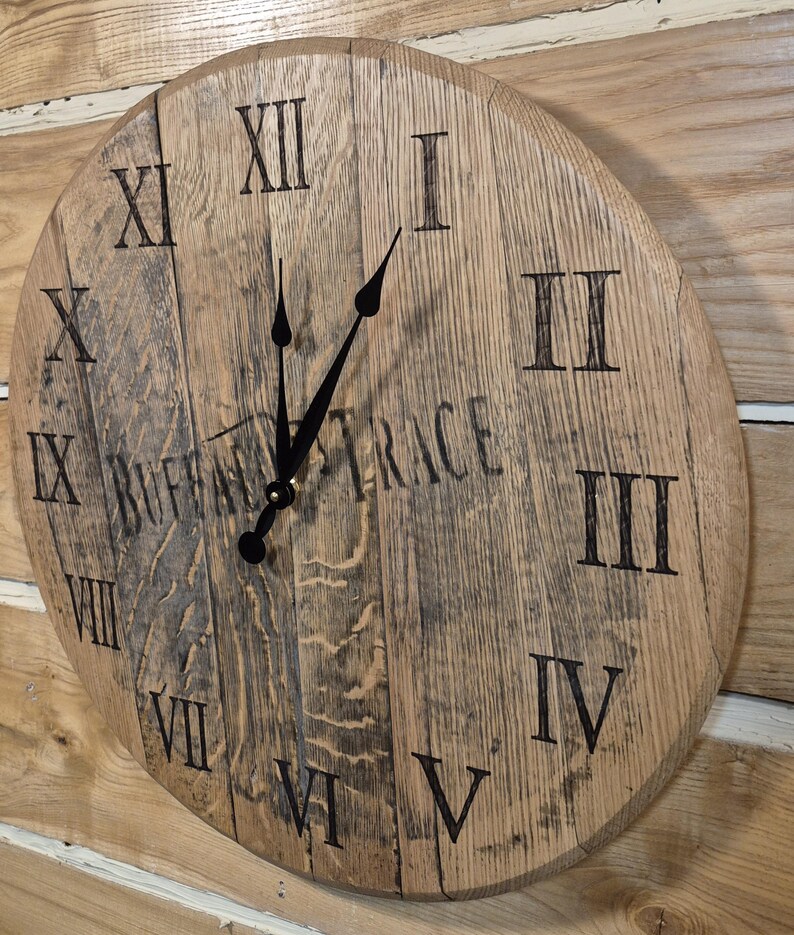 May include: A round wooden clock with Roman numerals and black clock hands. The clock face is made from reclaimed wood and features the text "BUFFALO TRACE". The clock is mounted on a wooden wall.