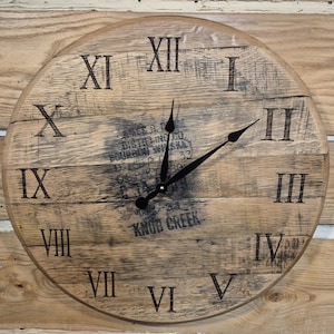 May include: Round wooden wall clock with black Roman numerals and black clock hands. The clock face has a distressed wood grain appearance with the text "KNOB CREEK" and other text. The clock is mounted on a wooden wall.