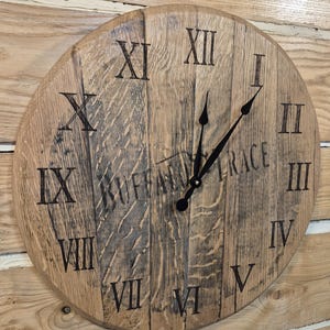 May include: A round wooden clock with Roman numerals and black hands. The clock face is made of light brown wood with visible grain and the text "BUFFALO TRACE" is partially visible. The clock is mounted on a wooden wall.