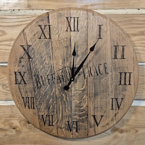 May include: Round wooden clock with Roman numerals and black clock hands. The clock face is made of wooden planks and features the text "BUFFALO TRACE". The clock is mounted on a wooden background.