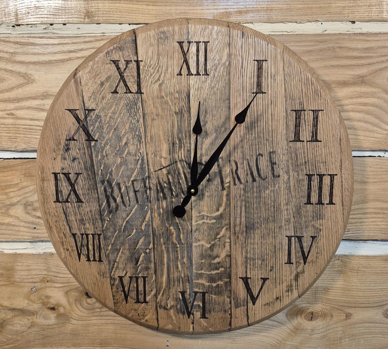 May include: Round wooden clock with Roman numerals and black clock hands. The clock face is made of wooden planks and features the text "BUFFALO TRACE". The clock is mounted on a wooden background.