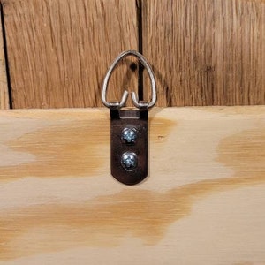 May include: A metal picture hanger with a D-ring and two screws, mounted on a light-colored wooden surface. The wood grain is visible, and the hanger is centered in the frame.