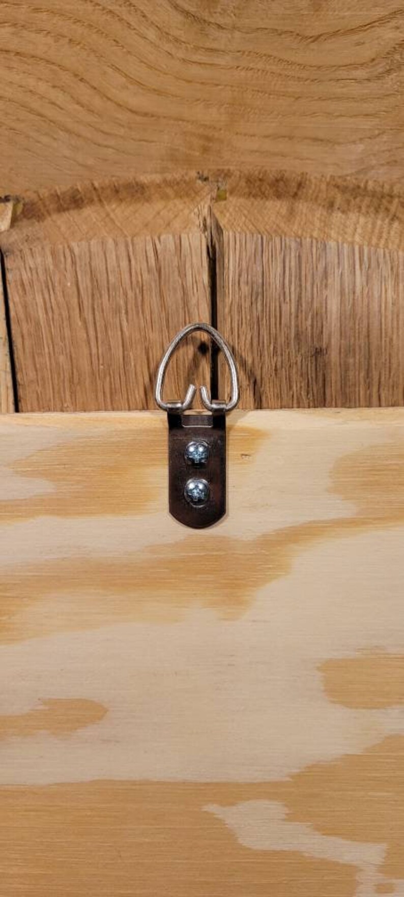 May include: A metal picture hanger with a D-ring and two screws, mounted on a light-colored wooden surface. The wood grain is visible, and the hanger is centered in the frame.
