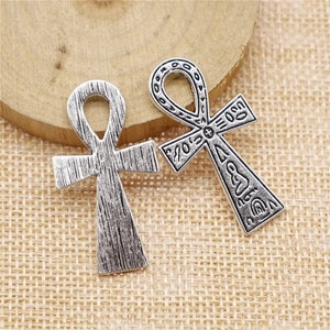 May include: Two silver-toned metal charms in the shape of an Ankh, an ancient Egyptian symbol of life. Both charms have intricate details and are engraved with symbols.