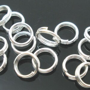 May include: A group of silver jump rings, a type of jewelry finding, are scattered on a black surface. The rings are all the same size and are made of a smooth, shiny metal.