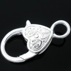 May include: Silver-toned heart-shaped clasp with a decorative design. The clasp has a round ring on one side and a hook on the other.