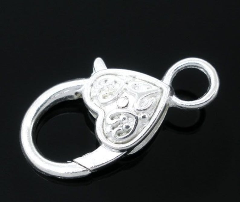 May include: Silver-toned heart-shaped clasp with a decorative design. The clasp has a round ring on one side and a hook on the other.