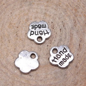 May include: Three silver metal flower-shaped charms with a hole for stringing. Two charms have the text "Hand made" on them. One charm is blank.