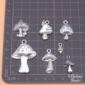May include: A set of eight silver mushroom charms with various designs, including a mushroom with a ladybug on top, a mushroom with white dots, and a mushroom with a textured surface. The charms are arranged on a gridded surface with a ruler in the background.