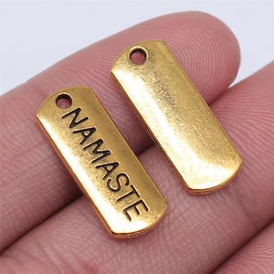 May include: Two gold-colored metal charms, one with the word "Namaste" engraved in black lettering, and the other plain.