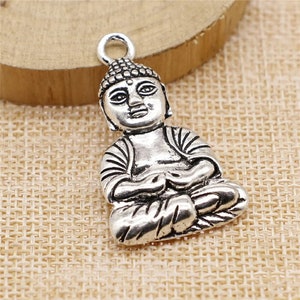 May include: Silver charm featuring a detailed depiction of a seated Buddha. The charm has a loop at the top for attaching to a necklace or bracelet.
