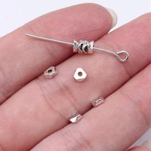 May include: Silver metal beads in various shapes, including triangular and square, with a hole in the center for stringing. The beads are displayed on a hand, with a few beads strung on a wire.