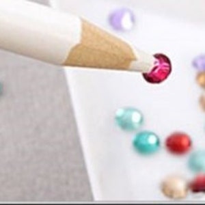 May include: A white tool with a wooden tip is used to pick up a small, red, round rhinestone from a tray of assorted rhinestones. The tray is white and triangular.