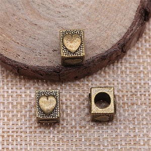 May include: Three antique gold-toned metal beads with a heart design. The beads are square shaped with a hole in the center for stringing.