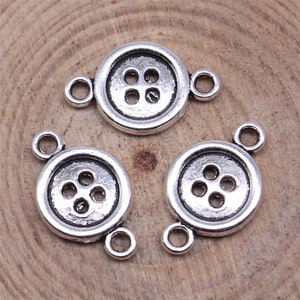 May include: Three silver-toned metal button charms with two jump rings on each charm. The buttons have a raised design with four holes.