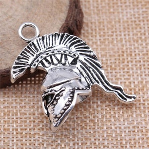 Op de afbeelding: Zilverkleurige metalen bedel in de vorm van een Spartaanse helm. De helm heeft gedetailleerde ribbels, een kam en een lus om aan een ketting of armband te bevestigen. De bedel heeft een antieke afwerking.