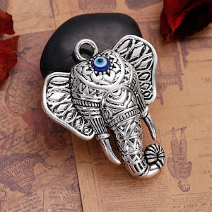 May include: A silver-colored elephant head pendant with intricate etched details. The elephant has a blue and white eye in the center of its forehead. The pendant is on a black stone, with a brown background.