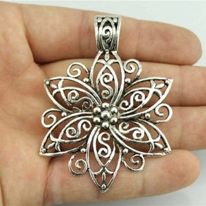May include: Silver-toned filigree flower pendant. The pendant features an openwork design with swirling patterns and a central cluster of small beads. The bail is a simple, rectangular shape. The pendant is held in a hand.