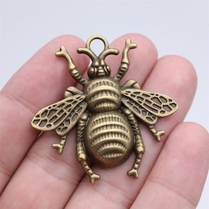May include: A gold-toned metal bee charm with detailed wings and a textured body. The charm has a loop at the top for attaching to jewelry or other crafts.