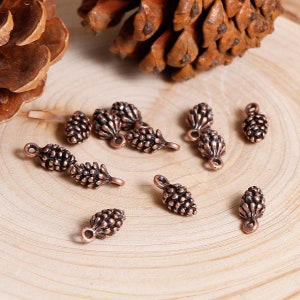 May include: A collection of 10 copper-colored pine cone charms with a loop for hanging. The charms are arranged on a wooden surface.