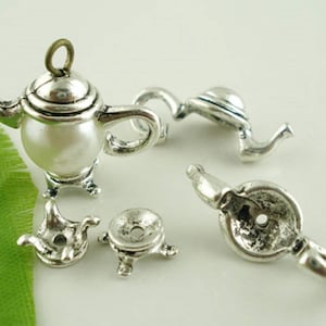 May include: Silver teapot charm with a pearl-like center and three legs. The charm is attached to a silver chain. There are also three other silver charms in the image, including a teacup and saucer charm.