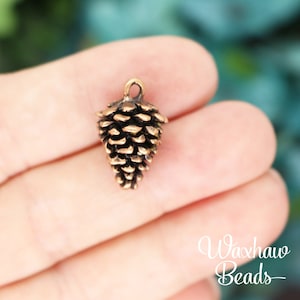 May include: A gold-toned metal pine cone charm with a loop for hanging. The charm is about 1 inch tall and is perfect for adding a touch of nature to jewelry or crafts.
