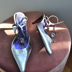 Peut inclure: Une paire de chaussures argent&eacute;es &agrave; talons hauts avec des brides &agrave; la cheville et un embellissement en pierres pr&eacute;cieuses roses. Les chaussures sont sur une surface en velours rose.