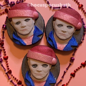 May include: Three round, gray-rimmed bath bombs featuring a masked face wearing a Santa hat and blue jacket. The bath bombs are arranged on a pink surface, with decorative beaded necklaces in the background. The text "@hocuspocusbath" is visible.
