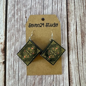 May include: A pair of dangle earrings featuring miniature book charms with the words "Fairy Tale" on the front. The charms are black with gold accents and are attached to silver hooks.