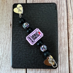 Puede incluir: Un marcapáginas de cuentas con estampado en blanco y negro con un dije de boleto morado que dice "Late Night Book Club" y "Admit One". El marcapáginas está unido a un lector electrónico negro con clips en forma de corazón plateados.