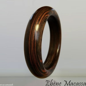 May include: A wooden bangle bracelet with a dark brown and reddish-orange striped pattern. The bracelet is smooth and polished, with a rounded shape. The text "Ebène Macassar" is visible at the bottom right.
