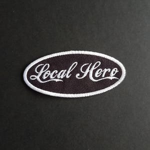 May include: A black oval embroidered patch with white stitching. The patch says "Local Hero" in a white cursive font.