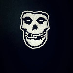 Op de afbeelding: Witte geborduurde patch van een glimlachende schedel met een zwarte omtrek. De schedel heeft twee oogkassen en een brede, tanden glimlach.